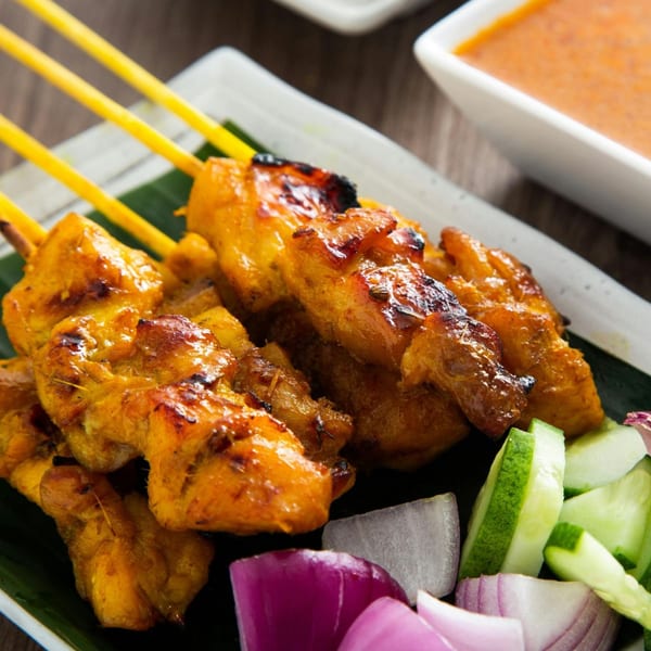 Grilled chicken satay skewers with a side of sliced red onions and cucumbers, and a dipping sauce.