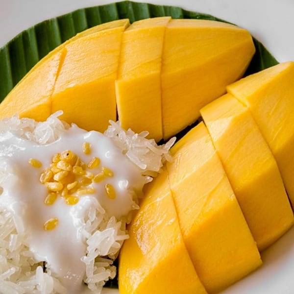 Close-up of mango sticky rice with coconut cream and golden crispy mung beans on a banana leaf.